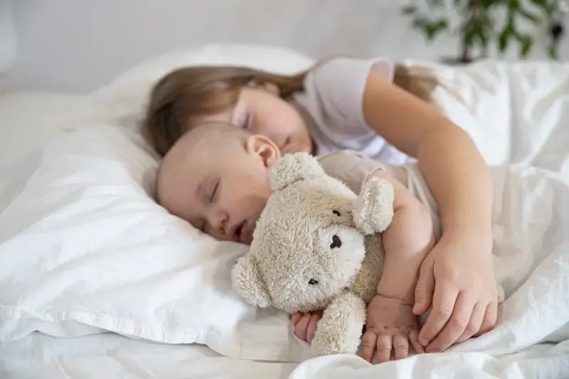Dzieci śpią wtulone w misia. Starsza siostra czuwa nad maluszkiem, pokazując, ile śpi dwulatek i jak ważny jest sen dla rozwoju.